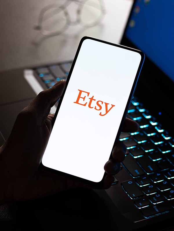 An open laptop on a desk with a pair of glasses on the side of it. A hand holding a cellphone and the words Etsy on the screen, and below the image is a blue box with text that states Starting and Running a Business on Etsy.